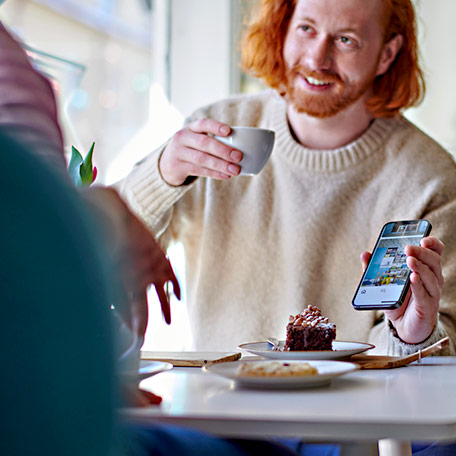 A man with red hair smiles holding a coffee cup and showing his phone. A slice of chocolate cake sits on the table in front of him.