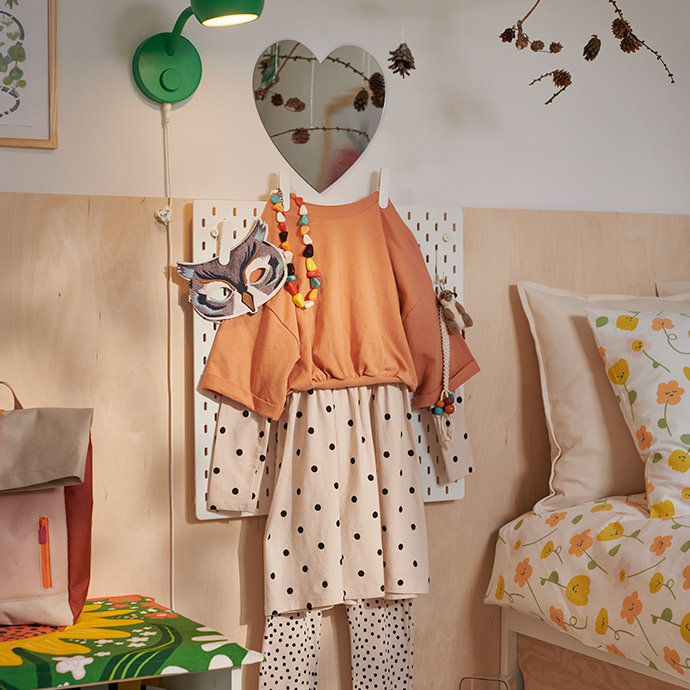 Un conjunto infantil de color beige y naranja cuelga de un panel perforado SKÅDIS, coronado por un espejo con forma de corazón HOLMÖARNA.