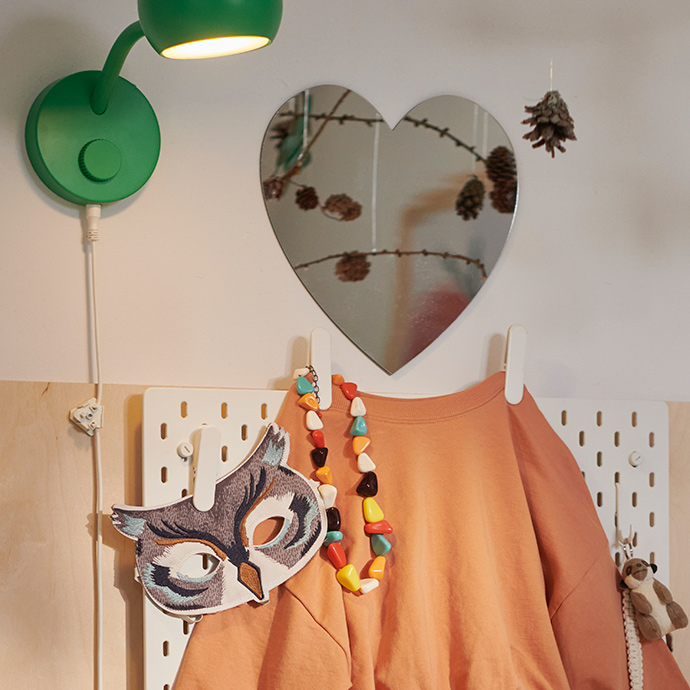 Un conjunto infantil de color beige y naranja cuelga de un panel perforado SKÅDIS, coronado por un espejo con forma de corazón HOLMÖARNA.