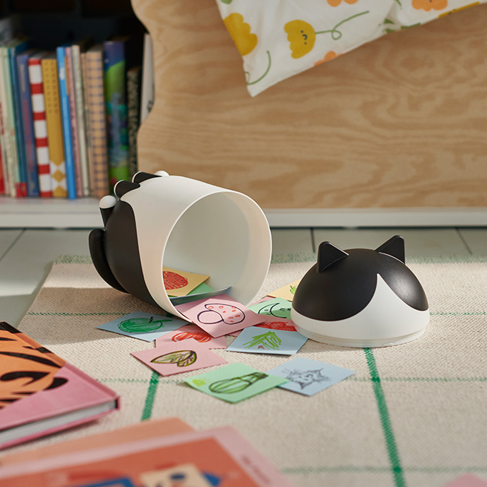 A cat-shaped GREJSIMOJS container lies opened on a carpet, revealing its content: a child’s paper cards.