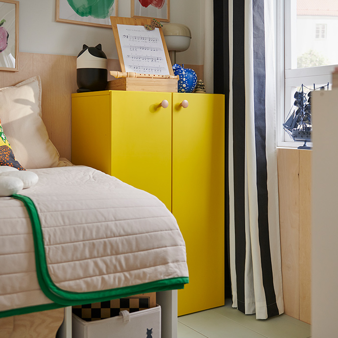 Una combinación de almacenamiento LASTARE de color amarillo brillante, con juguetes infantiles sobre la parte superior, se encuentra junto a una cama con una colcha blanca y verde.