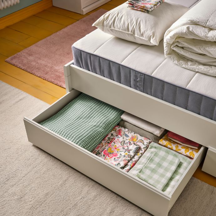 A bedroom with an off-white FÅGELFJÄLLET bed frame and four storage boxes, one filled with folded bedding.