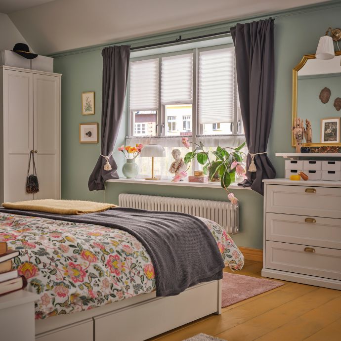 A bedroom with a bed dressed in floral bedding and a BEKVÄM stool by the window. KORGMOTT blackout curtains frame the window.