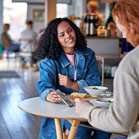 A smiling woman in a denim jacket shows her phone screen to a friend while sharing a meal at a bright restaurant table.