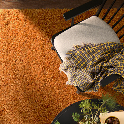 Una alfombra naranja ÄRENDE sobre un suelo de madera con una silla negra y una manta drapeada. La alfombra es grande, suave y lujosa, proporcionando calidez y comodidad.