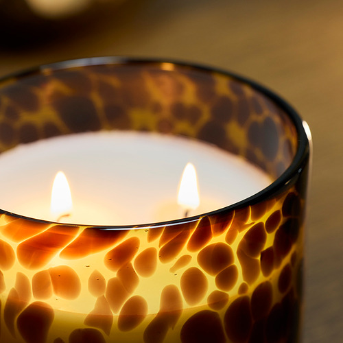 Una vela encendida en un recipiente de cristal estampado, que emite un resplandor cálido y acogedor con tres mechas, mostrando una artesanía de alta calidad.