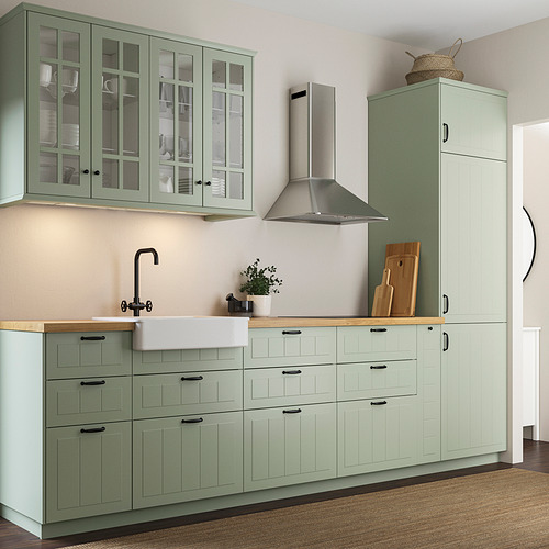 Cocina moderna con gabinetes verdes, encimera de madera, campana de acero inoxidable, fregadero blanco, manijas negras, planta, tablas de cortar, despensa alta, tapete marrón.