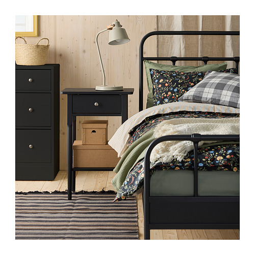 Dormitorio con cama de metal, manta verde oscuro, almohada floral, mesita de noche negra, lámpara y alfombra a rayas.