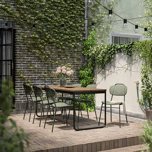 Comedor al aire libre con una mesa extensible de acacia y sillas acolchadas verdes, rodeado de ladrillo y vegetación.