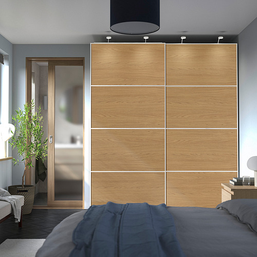 Dormitorio moderno con gran armario de madera MEHAMN . Los armarios tienen laterales de roble claro deslizándose para acceso. Cama gris suave, plantas y suelo gris-marrón.