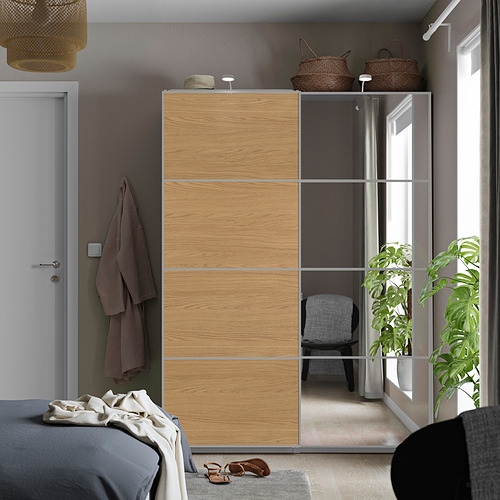 Dormitorio moderno con armario de madera clara, acabado de espejo, puerta blanca, ropa de cama gris, cestas y plantas.