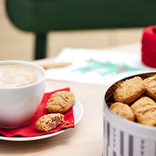 Imagina una taza blanca de café espumoso en una servilleta roja, con galletas en un plato, y una lata de galletas al lado.