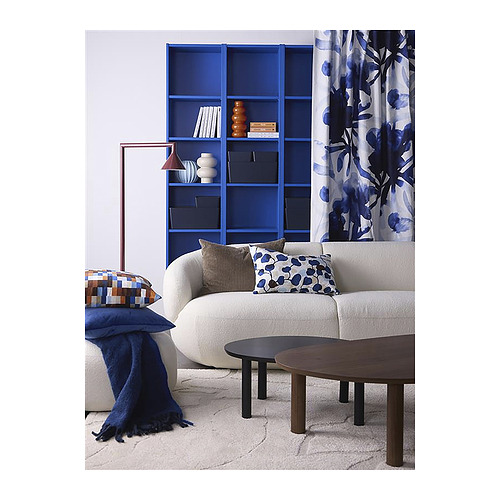 Un salón acogedor con sofá blanco, cojines de colores, estanterías azules, mesas de madera y cortinas ornamentadas con hojas estampadas.