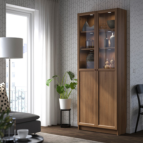 Elegante librería BILLY de madera con puertas de vidrio en la sala de estar, al lado de una ventana, sosteniendo jarrones, sofá gris en contraste, tonos cálidos.
