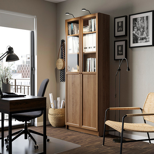 Estantería con puertas de vidrio cerca de la ventana, libros, decoración, escritorio, lámpara, cuadros. Bien iluminado, organizado.