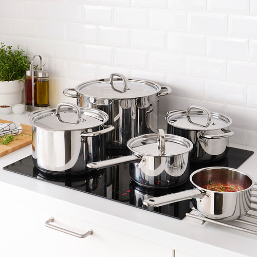 Un juego de ollas y sartenes de acero inoxidable IKEA 365+ sobre una estufa negra. Las ollas tienen tapas y se utilizan para cocinar diversos alimentos.