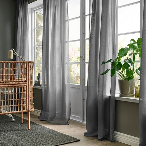 Habitación moderna con cortinas grises transparentes STENFRÖ, planta grande en maceta, silla de mimbre. Las cortinas suavizan la luz, se encharcan en el suelo, enmarcadas por ventanas altas.