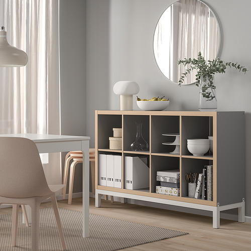 Aparador KALLAX gris claro con detalles en madera, que sostiene jarrones, cuencos y plantas, contra una pared gris claro debajo de un espejo redondo.