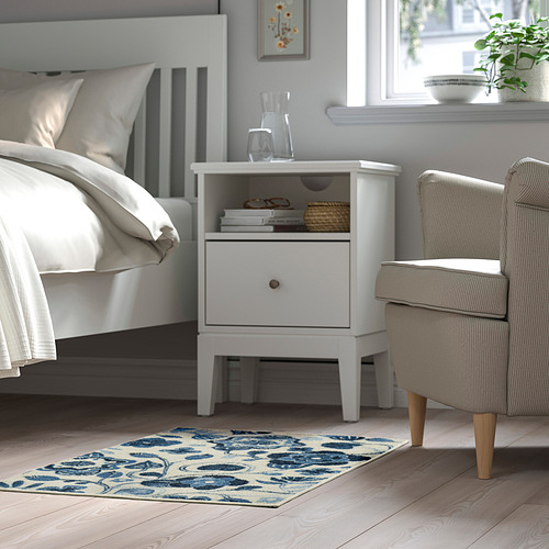 Un dormitorio con una alfombra blanca KANTLINJE, azul y blanca, suave bajo los pies, colocada junto a una cama y una silla, que añade calidez y confort a la habitación.