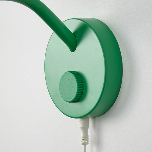 Lámpara verde de sobremesa montada en una pared blanca, con base circular y mástil ajustable. Incluye un control de dial y está conectado mediante un enchufe.