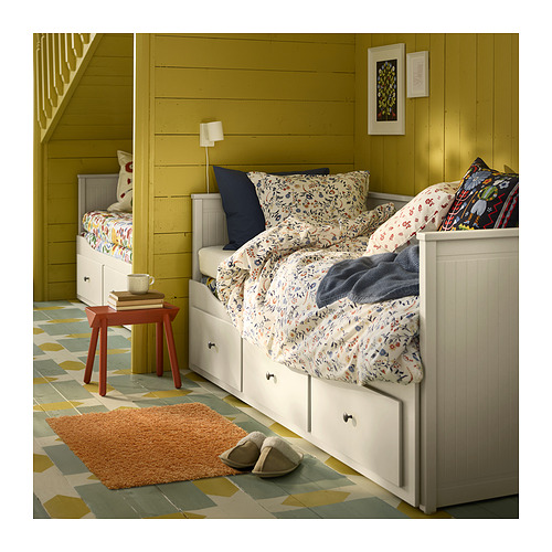 Un dormitorio acogedor con paredes verde oliva, camas blancas, ropa de cama floreal, una mesita de madera y una suave alfombra marrón sobre un suelo geométrico de azulejos de colores.