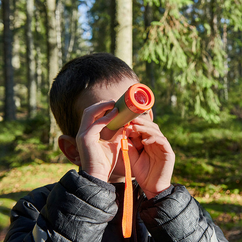 Un niño sostiene un telescopio naranja, explorando la naturaleza en una zona boscosa. El telescopio ayuda a observar objetos distantes, fomentando la curiosidad al aire libre.
