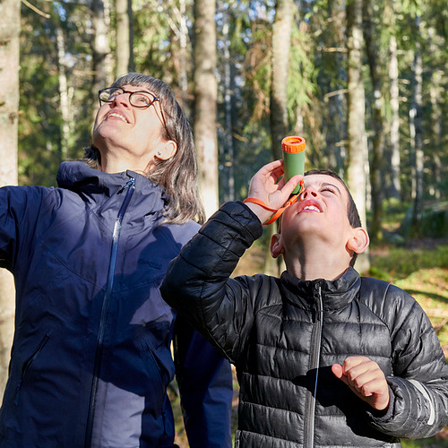 Dos personas en un bosque, una sostiene a un niño usando un pequeño telescopio verde y naranja, mirando hacia arriba.