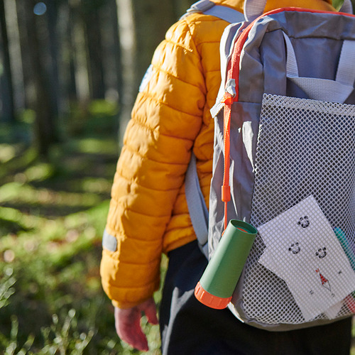 Un niño que lleva una chaqueta naranja lleva una mochila con una botella de agua y un bloc de notas en un entorno al aire libre.