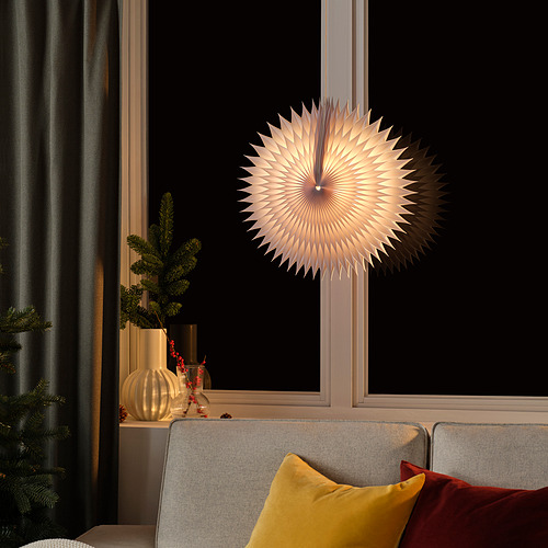 Lámpara STRÅLA: blanca, en forma de estrella, cuelga de la ventana, ilumina la acogedora sala de estar. Sofá gris con almohadas amarillas y rojas debajo, vegetación al lado.