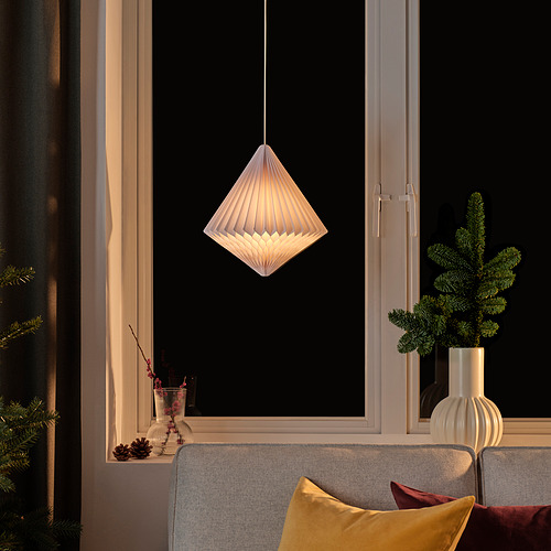 La lámpara STRÅLA cuelga, luz cálida. Sofá gris con almohadas, planta cerca, encima de la ventana.