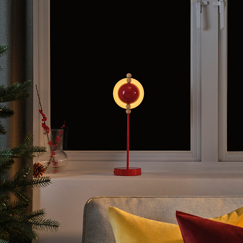 La lámpara roja STRÅLA, de diseño moderno, se encuentra en el alféizar de la ventana, reflejándose en el vidrio, rodeada de una decoración festiva.