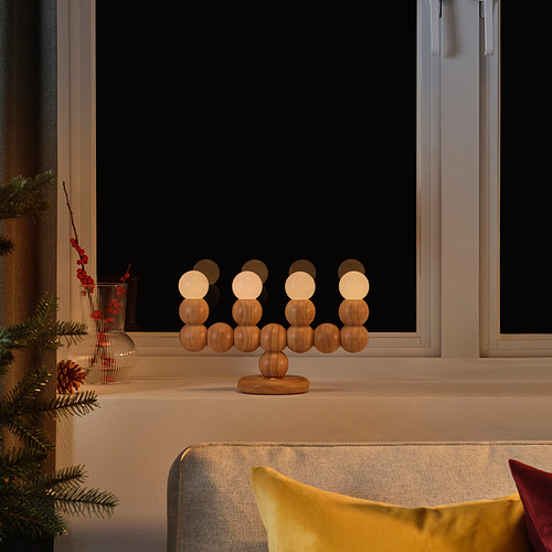 La lámpara STRÅLA brilla suavemente en el alféizar de la ventana con decoración navideña, de madera y esférica, junto a un cojín amarillo.