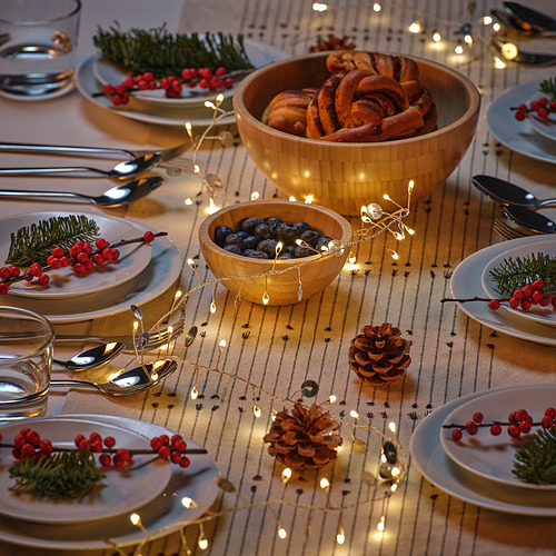 Juego de mesa navideña con platos blancos, bayas, piñas, luces led de disco de plata STRÅLA que añaden un cálido resplandor, cuencos de madera con pan y arándanos.