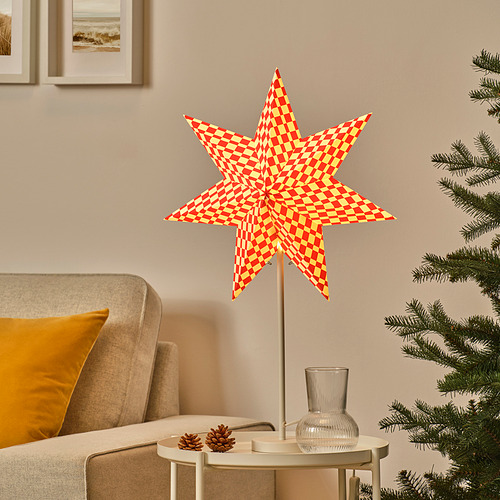 Lámpara en forma de estrella de la colección STRÅLA en rojo y amarillo sobre mesa junto al árbol de Navidad.