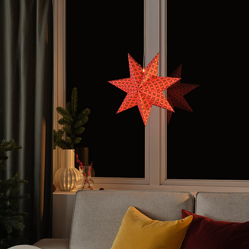 La decoración de estrella de papel rojo cuelga fuera de la ventana oscura, reflejando luces, cerca del sofá gris con almohada amarilla, creando un ambiente acogedor.