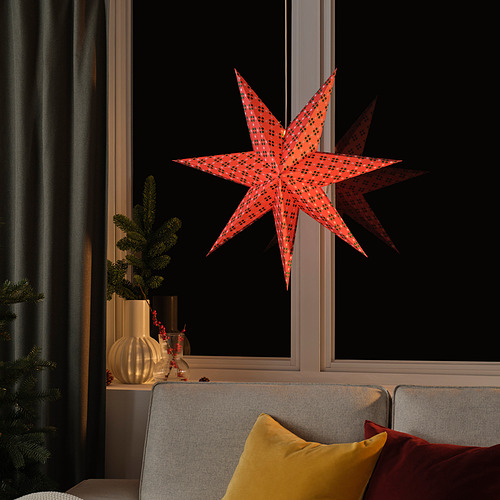 La decoración STRÅLA en forma de estrella de color rojo brillante cuelga grande en la ventana, rodeada de acogedores muebles de sala de estar.