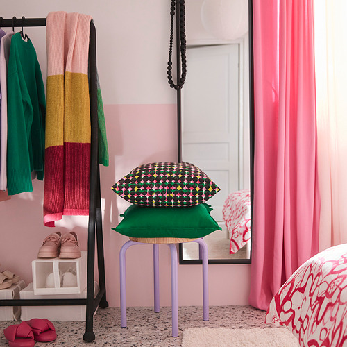 Cojín colorido sobre un taburete morado, rodeado de ropa y zapatos, decoración vibrante, cortinas rosas.