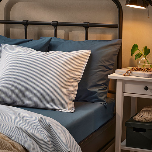Cama con almohadas azules y blancas, cabecero metálico, mesita de noche con lámpara y planta.