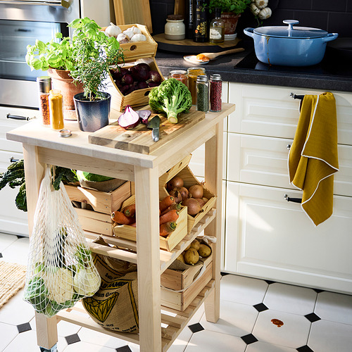 Isla de cocina de madera con cajas de verduras, hierbas en macetas y utensilios de cocina encima. Una toalla amarilla cuelga de un tirador de cajón.