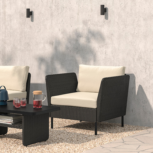 Moderno salón exterior de mimbre negro con cojines, mesa de centro, copas de vino y altavoz, apoyado contra una pared de hormigón gris.