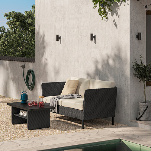 Patio moderno con juego de salón de mimbre VITTSKÄR negro. El sofá, los cojines y la mesa relájate cerca de la piscina. Pared gris, vegetación y suelo de guijarros.