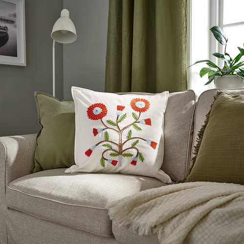 Cojín decorativo llamado FACELIOR flores de colores sobre algodón blanco. Colocado en un sofá con cortinas, es más pequeño que los cojines que lo rodean.