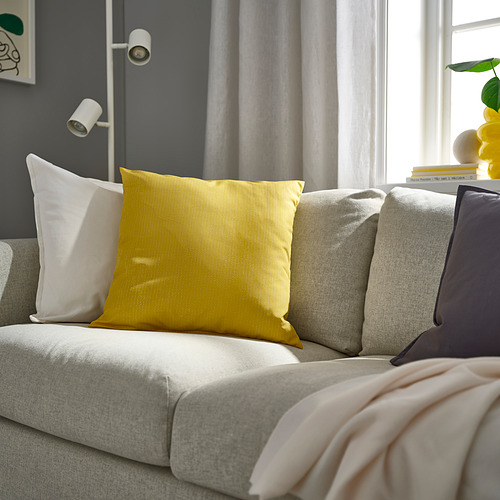 Sala de estar moderna con una gran almohada PROFETBLOMMA amarilla, sofá gris claro, almohadas blancas y moradas, manta rosa, cortinas grises y apliques de pared.
