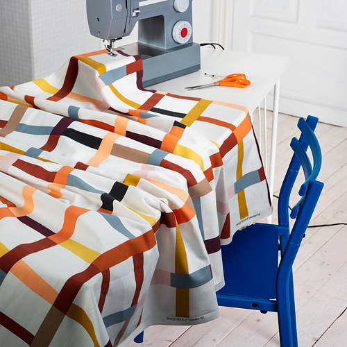 Una estación de costura con tejidos a rayas coloridas colgada sobre una mesa blanca, una máquina de coser y una silla azul, lista para crear cortinas o decorar el hogar.