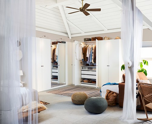 Armarios blancos luminosos y minimalistas para el dormitorio, ventilador de techo, pufs de tela afelpada y cortinas blancas transparentes que crean un ambiente sereno.