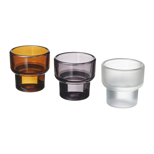 Tres coloridos portavelas cilíndricas de vidrio en marrón, gris y blanco están dispuestos uno al lado del otro, mostrando su diseño simple y elegante.