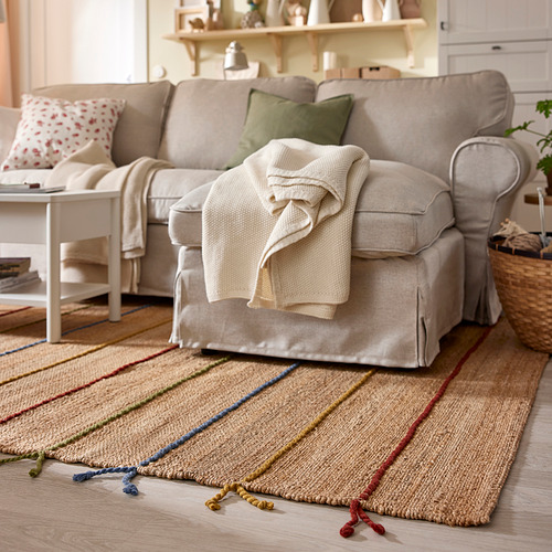 Alfombra de yute tejida a mano con rayas de colores y borlas de lana en la sala de estar, debajo del sofá beige y la mesa auxiliar.
