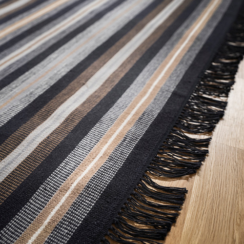 Alfombra debilitada de rayas negras y marrones con flecos sobre suelo de madera.