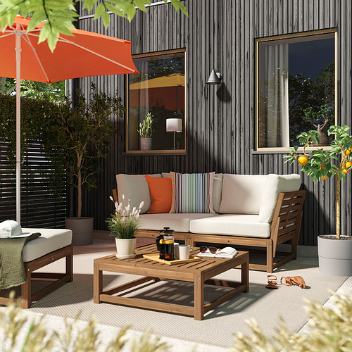 Acogedor patio exterior con sofá acolchado beige NÄMMARÖ, mesa de centro de madera, sombrilla naranja, plantas y árbol en maceta.