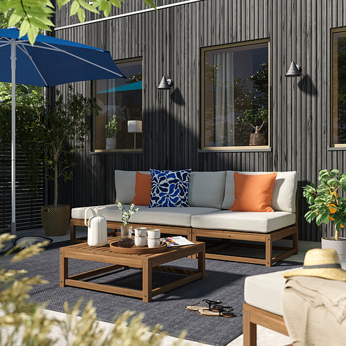Moderno patio al aire libre con sofá gris NÄMMARÖ, almohadas naranjas y azules, mesa de centro de madera, sombrilla azul, plantas y fondo de casa oscuro.